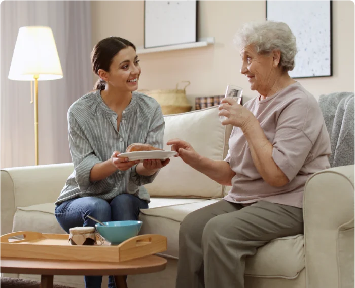 Young woman caregiver handing plate to smiling elderly woman on living room sofa.