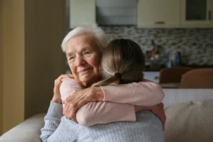 Elderly woman and young caregiver embracing warmly in sunlit living room with kitchen background.