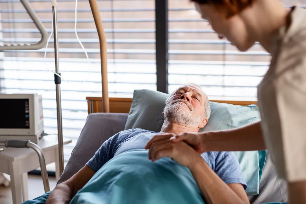 Nurse gently holding hand of elderly man resting in hospital bed with IV and monitor.