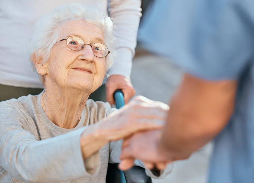 Nurse holding hand of smiling elderly woman with glasses and walker outdoors.