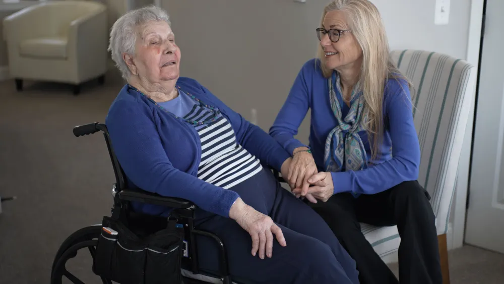 Caregiver holding hand of elderly woman in wheelchair beside striped chair in lounge area.
