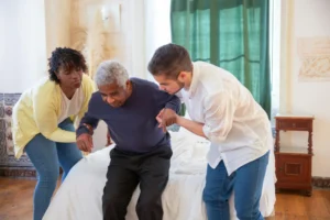African American woman and young man assisting elderly Black man rising from bedroom bed.