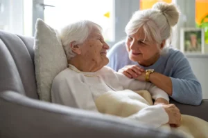 Elderly woman smiling while caregiver supports her from behind in a bright room