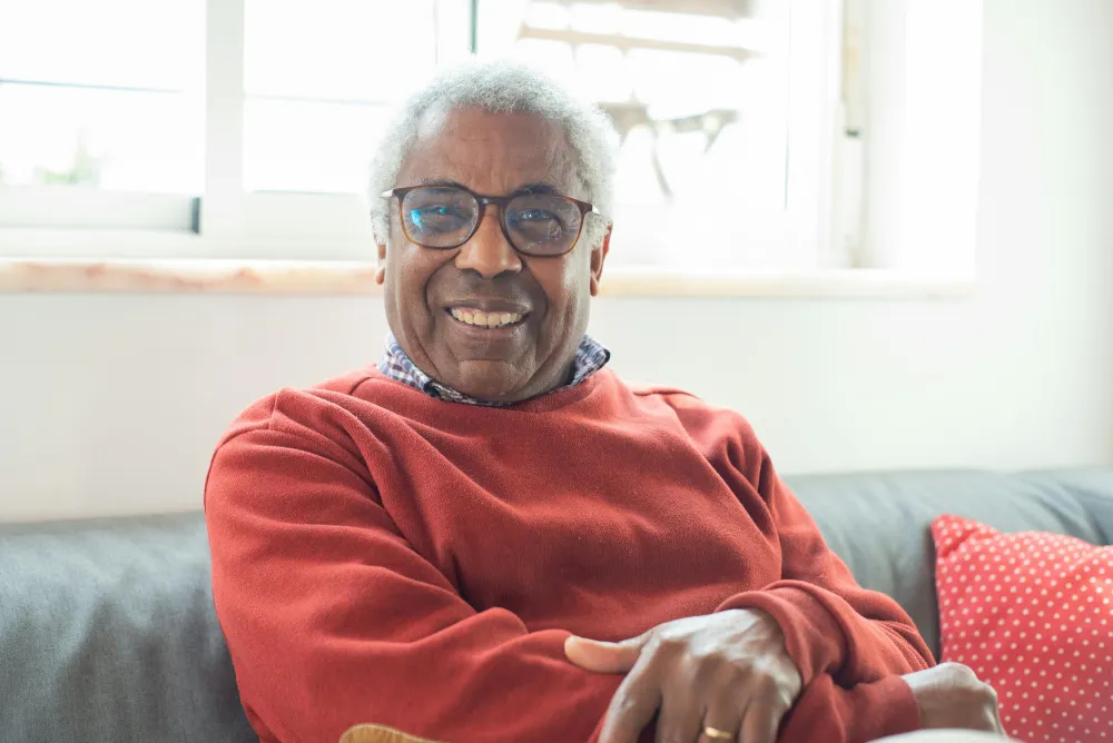 Elderly Black man with glasses smiling warmly while seated on gray sofa near sunlit window.