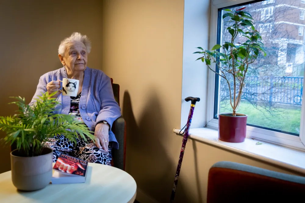 Elderly woman sitting by window holding a cat mug with a walking cane nearby