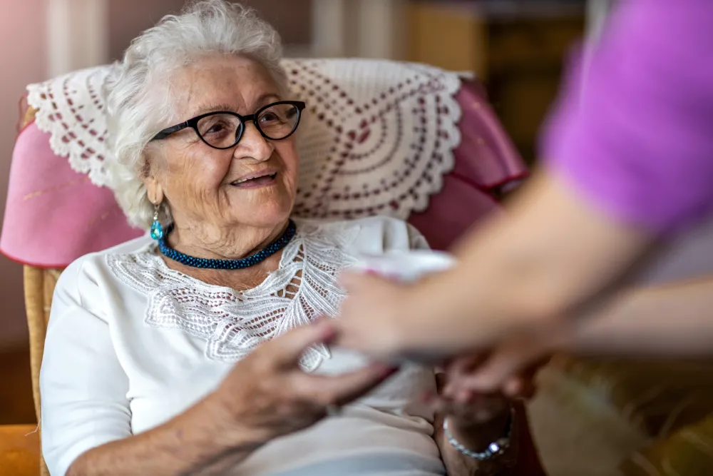 Elderly woman smiling and receiving a cup from a caregiver in a comfortable setting