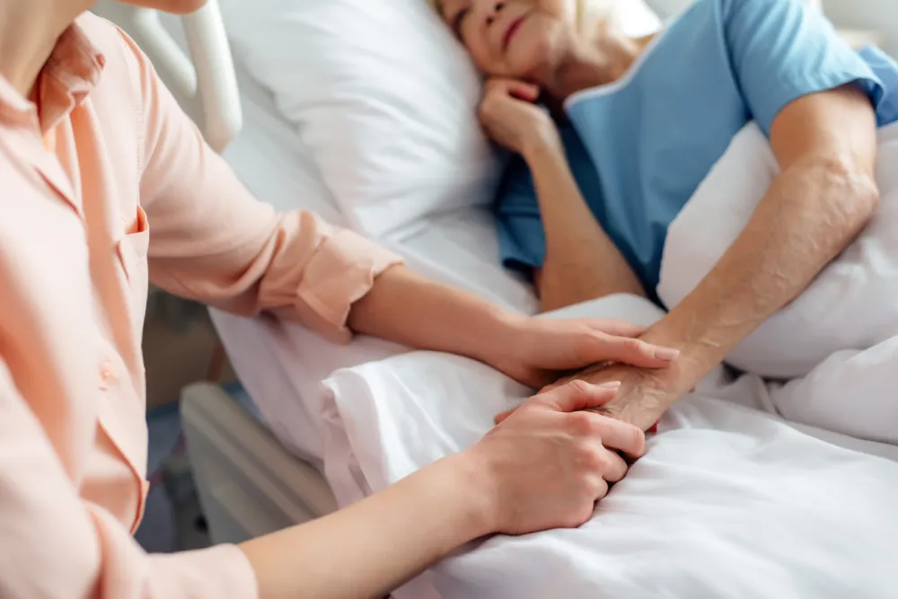 Caregiver holding elderly patient’s hand while they rest in a comfortable bed